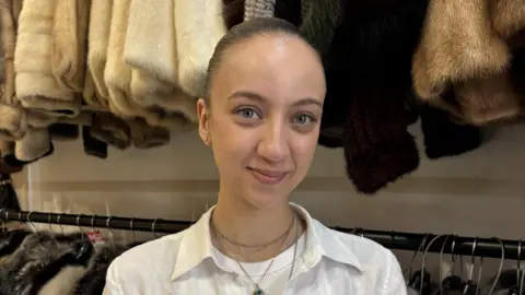 Mel Kaplan smiles at the camera. Visible behind her is a rack of fur coats in shades of white, cream, black and brown. Mel has her hair slicked-back into a ponytail and is wearing three layered necklaces and a satin shirt over a white T-shirt. It is a head and shoulders shot of her.