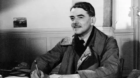A black and white photo of a man at a desk, holding a pencil, wearing a shirt, tie, jumper and a coat, with short black hair and a short black moustache.