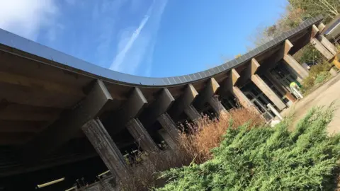 The sweeping frontage of the Eco Centre building, a modern building with wooden pillars and a curved roof. Plants are visible in front of the building, which has been photographed from an angle.