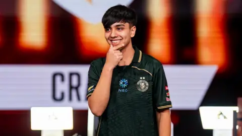 Esports Foundation Jaime "Craime" Bustos stands on-stage, smiling, his hand held up to his chin, as he is announced as the winner of the EWC's Street Fighter 6 contest.