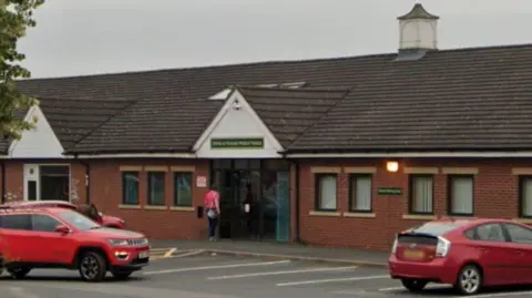 Google The front of a one-storey red brick building with two red cars outside and a green sign above the entranceway