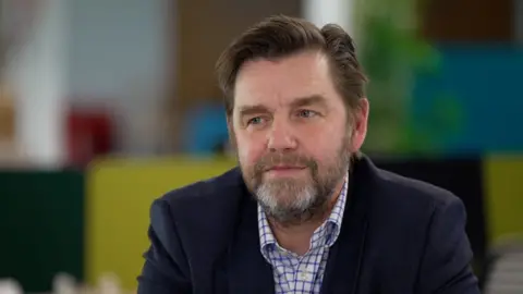 A portrait photo of Mayor Nik Johnson who has dark coiffed hair, a brown beard with a touch of grat on his chin. He is wearing a white shift with purple checks and has a dark blazer on. 