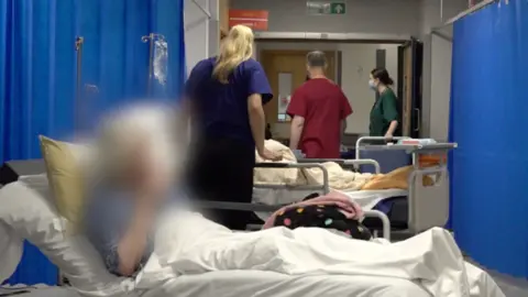 BBC A side ward at the A&E unit at Morriston Hospital Swansea, with medical staff attending to an unseen patient in the background, while another patient in bed - face blurred to protect his identity - waits.