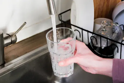 Hand holding a glass, filling it with water, with stacked dishes in background