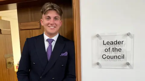 George Finch leans against a door frame. A sign to his left reads 'leader of the council'. He wears a dark suit, white shirt and purple tie.