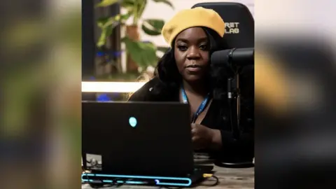 Jonas Kontoutas A woman wearing a yellow hat sits in a high-backed gaming chair at a desk. A streaming microphone is set up close to his face and in front of him is a black laptop with a glowing alien logo in the middle of the open lid.