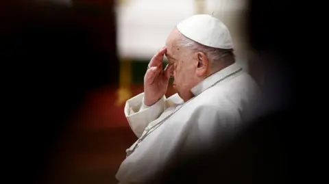 A picture of Pope Francis, taken from the side. He is touching his face.