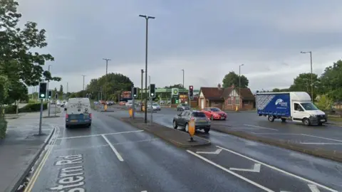 A busy junction on Eastern Road, with cars and vans travelling in both directions and an array of traffic lights. In the background there is the forecourt of a bp garage. 