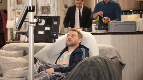 Coronation Street/ITV Actor Peter Ash portraying the character Paul Foreman in the ITV soap Coronation Street. The character is sat on a sofa in a kitchen staring at a screen.