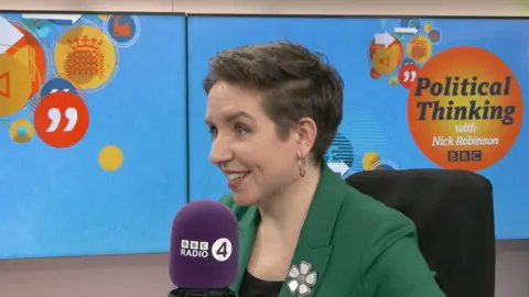 Carla Denyer, wearing a green suit, sitting and smiling in a radio studio in front of a BBC Radio 4 microphone