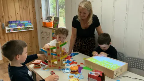 A woman with shoulder length blonde hair, a black short sleeved top and a leopard print skirt stands over a table where three young children are playing. One little boy is playing with some cars. A lttle girl is helping him. And another boy is playing with letters on a light box.