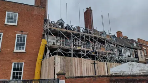 A black and white timbered building covered in scaffolding and surrounded by wooden fencing with a red brick building alongside