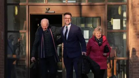 PA Media Charles Hanson pictured leaving the court with his parents. He is wearing a suit and tie. His dad is in a black puffer jacket with a scarf around his neck. His mum is in a red coat.