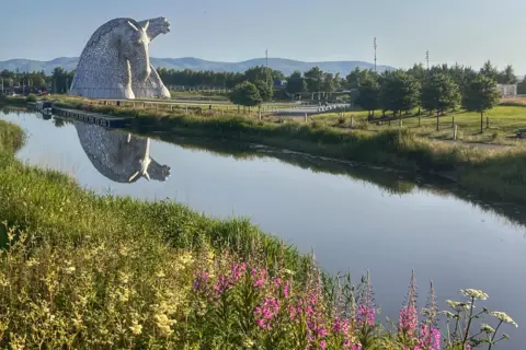 BBC Weather Watchers - Maggie D Large steel horse-head sculpture beside a calm canal, its reflection mirrored in the water, with green parkland, trees, and distant hills under a clear blue sky.