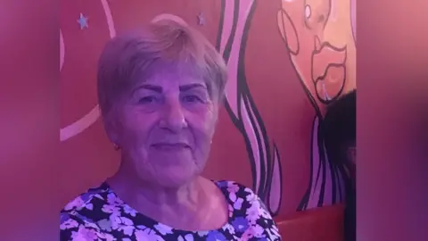 a woman in a floral blouse, with short grey-blonde hair, smiles at the camera