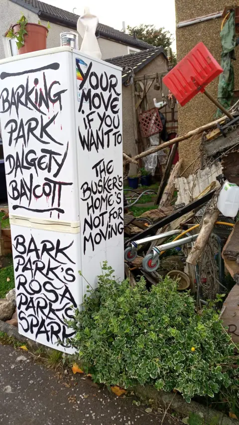 Denis Carbonaro A fridge in the front garden surrounded by piles of wood and other items. Text on the fridge reads BARKING PARK DALGETY BAYCOTT, BARK PARK'S ROSA PARKS SPARK, YOUR MOVE IF YOU WANT TO, THE BUSKER'S HOME'S NOT FOR MOVING in black.