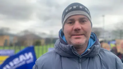 Middle-aged man with short grey stubble wears a wooly hat and puffer jacket and is standing outside. In the background of the photo union flags can be seen.