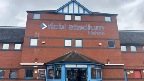 A large red-bricked building with the words DCBL Stadium Halton on the front. 