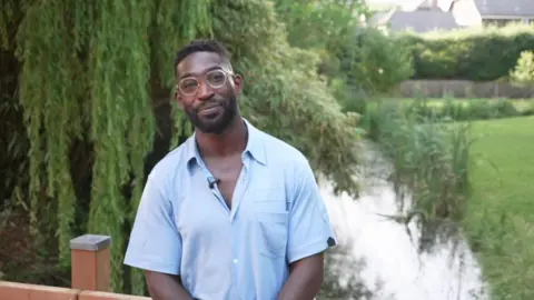 Tinie Tempah in a blue shirt and glasses smiling at the camera while on a bridge over a river in a green area.