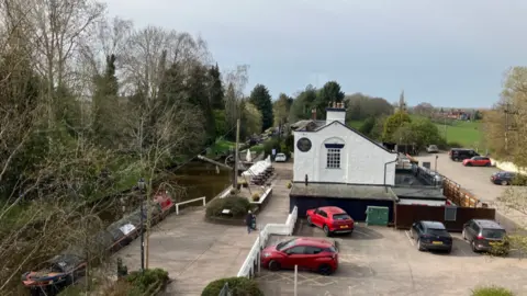 Looking down on a large white building alongside a canal, with a barge moored at the side. There are cars parked to the side and back of the building and there is greenery all around.