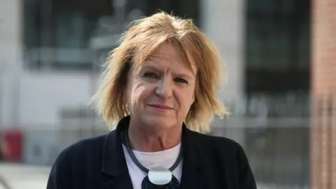 Head and shoulders photo of Deborah Coles. She has straight blonde shoulder-length hair with a centre parting. She is wearing a dark jacket with a white top underneath, and a large black and silver necklace. She is standing in front of a building with railings outside, and is looking directly into the camera