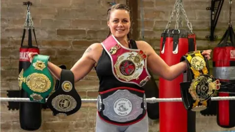 Michelle Minaides standing holding five boxing belts smiling at the camera inside a boxing gym.