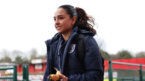 As imagens Getty estão sorrindo com uma banana na mão. Ele tem uma jaqueta de inverno da Marinha. No fundo, você pode ver um campo de futebol.