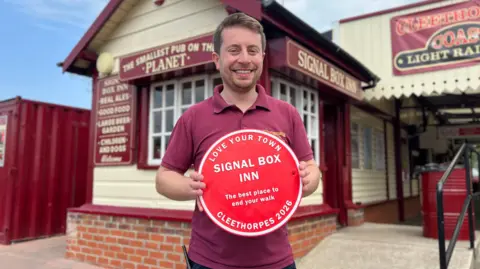 BBC A man, wearing a burgundy polo shirt, smiles holding a bright red circular plaque that reads: 'Love Your Town, Signal Box Inn, the best place to end your walk, Cleethorpes 2026'. In the background is a cream building called 'The Smallest Pub on the Planet'.