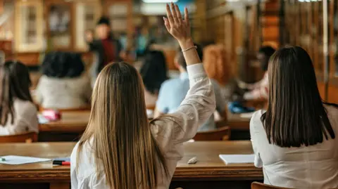 Students sitting in a classroom. The photo is taken from behind them. One of the pupils has their hand raised.