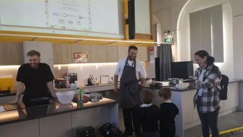 Northamptonshire Community Foundation Two young children and woman at a counter talking to man with an apron on. The woman and the man are smiling and another man is to the side also smiling.