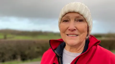 Older woman in a red jacket and white hat in front of a hedgerow