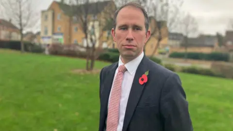 Thames Valley Police and Crime Commissioner Matthew Barber photographed outside on an overcast day. He is looking away from the camera. He has a paper poppy on his lapel.