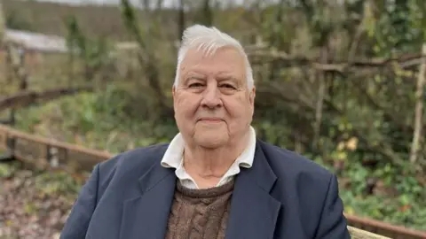 George Carden/BBC An elderly man with white hair, smiling as he looks at the camera. He is wearing a white shirt with brown knitted jumper and dark blue blazer. He is stood in front of a blurred brown fence with green hedges and trees in the background