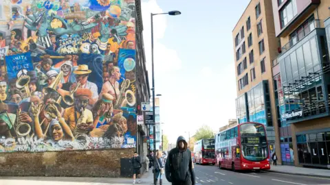 A mural on a wall with colourful drawings of musicians and local people. It stands opposite a modern Hackney council building. 