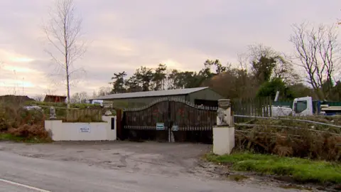 White pillars sit either side of black gates and on the white pillars are a pair of lions. Behind there is a large green shed and a lorry. Also visible is a white van.