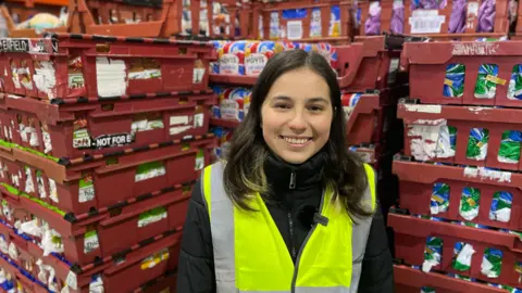 Isabella, a young woman with dark shoulder-length hair. She is wearing a luminous jacket and is stood in front of some stacked shelves packed with loaves of bread.