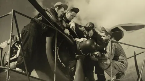 Historic England Archive A black and white still image from a film, which shows airmen around the propeller of an aircraft.