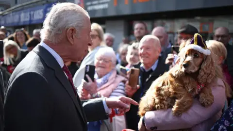 PA Media King Charles, a man with grey hair , is dressed in a suit and is greeted by a woman in a pink coat holding a red coloured cocker spaniel who is wearing a knitted crown. 
A number of people are in the background taking photos. 