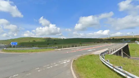 Google Junction 37. It is a two-lane road with a blue sign which reads 'The South M6'. There are cars in the distance and two wind turbines on the hills to the right.
