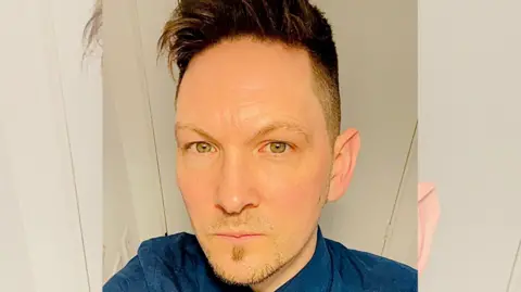 Supplied John Payne takes a selfie from a raised angle. He is wearing a blue shirt and is standing in front of a white door. He has green eyes and dark hair, styled in a quiff. 