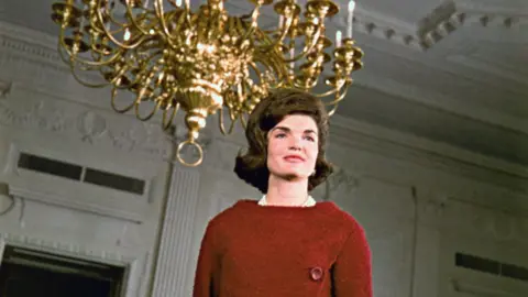 Jackie Kennedy wearing a red dress stand beneath a gold chandelier