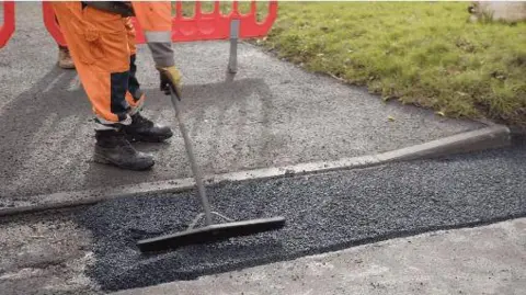 A person is working on repairing a road. They are visible from the hips down, wearing high visibility orange clothing, yellow gloves and dark work boots. There is a bright orange temporary barrier behind them, and they're using a long handled tool to level out the new tarmac that has been laid. There is a grassy area to the right of the works.