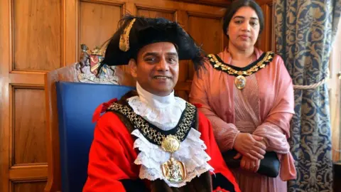 A man in black and red mayoral robes and chains of office sits in a ceremonial chair. Standing behind him is a lady in a pink dress, who also wears mayoral chains.