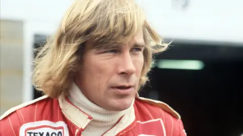 A head-and-shoulders colour photo of a man in his late twenties, with long blonde hair. He is looking away from the camera to the right of frame, wearing motor racing overalls with the logo of sponsors Texaco just visible near his right shoulder.