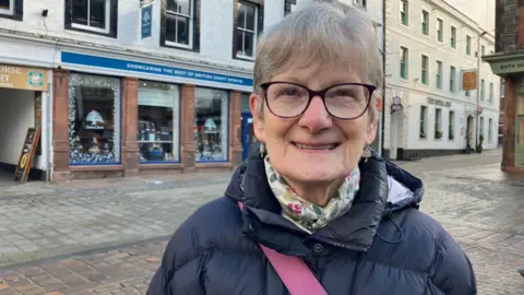 A head-and-shoulder image of Jenny Stokes. She. She has short grey hair and wears glasses with a dark rim. She is wearing a blue puffer jacket with a white neckerchief with a pastel pattern. She is standing in the pedestrian area in Keswick.