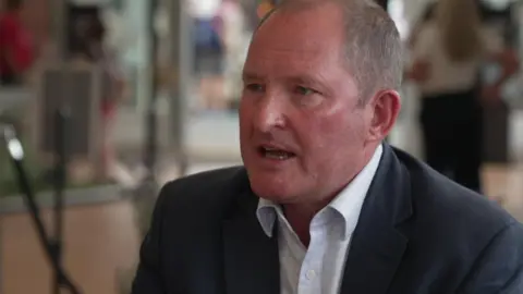 Steve McLean being interviewed with a blurred background behind him and wearing a dark suit and light shirt open at the neck.