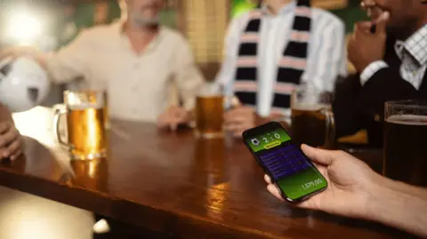 A person holding a phone which shows a generic football score on the screen alongside other statistics. They are sat at a table with pints of beer on it. Three blurred men sit on the other side of the table.
