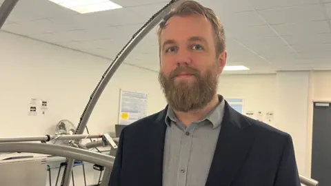 A man with a beard, a grey shirt and a blue jacket stands in a laboratory facing the camera.