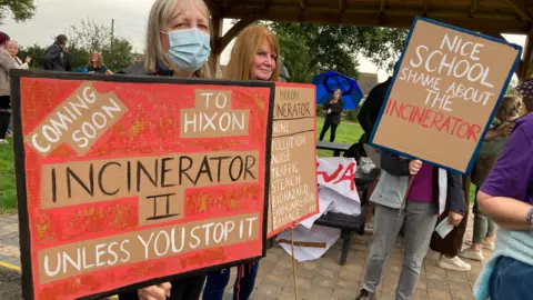 BBC Residents gather at a protest. Some are holding placards and signs opposing the plans for the clinical waste incinerator. 