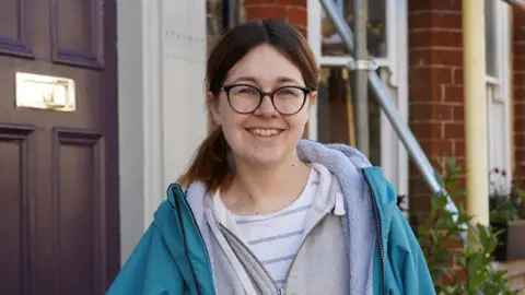 Jade is wearing a grey hoodie and blue coat, with black glasses, she is standing in front of a brown doorway, smiling.